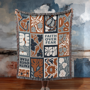 Decorative blanket with floral patterns and 'Faith Over Fear' text, held by a person against a textured wall.