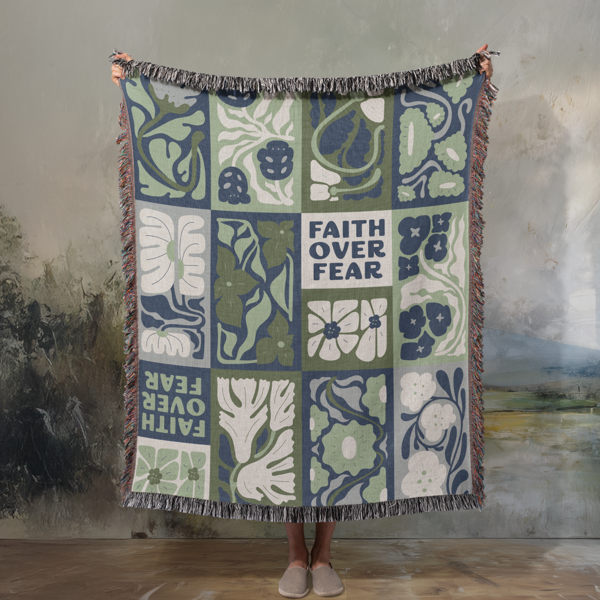 Decorative throw blanket with floral patterns and 'Faith Over Fear' text, held by a person against a textured wall.