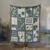Decorative throw blanket with floral patterns and 'Faith Over Fear' text, held by a person against a textured wall.