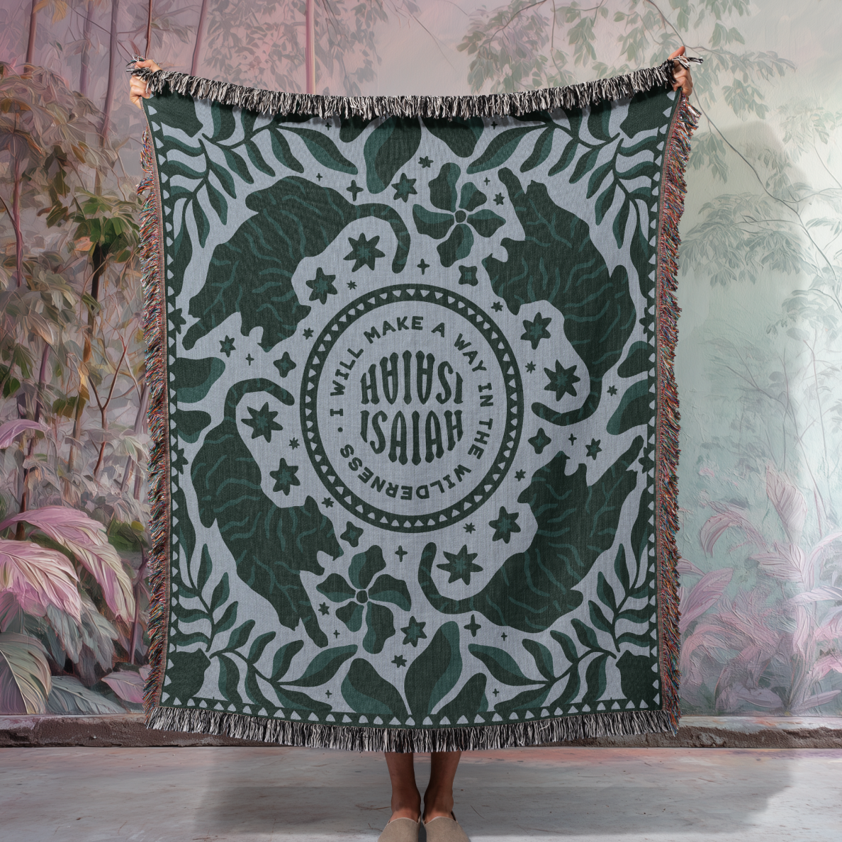 Green and white patterned throw blanket with text, held by a person in a room with plants.