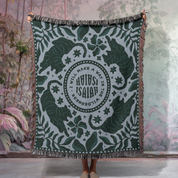 Green and white patterned throw blanket with text, held by a person in a room with plants.