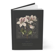 A hardcover journal or book with a dark charcoal-gray cover, featuring a vintage botanical illustration of white flowers with green leaves and a gray ornamental border. The title 'JOHN 1:18' and a subtitle are printed in light gray text below the illustration.