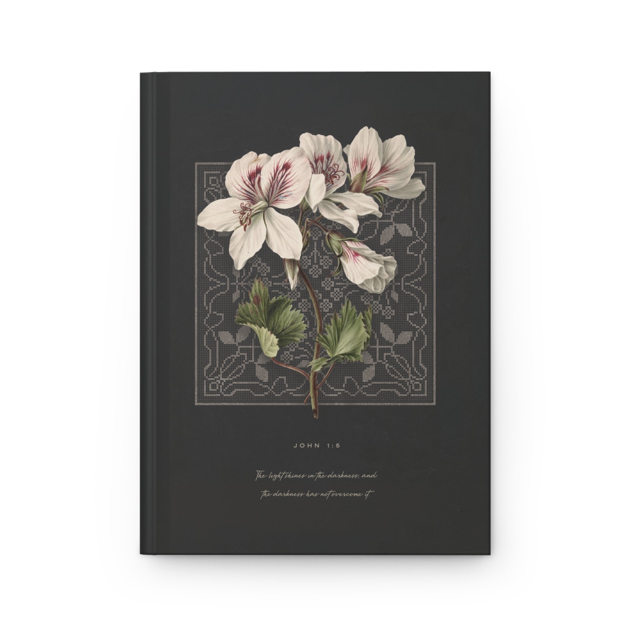 A dark charcoal gray hardcover journal featuring a vintage botanical illustration of white flowers with green leaves, framed by an ornate border, with the bible verse John 1:9 and a scripture printed below.