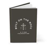 Black notebook with 'Built on the Rock, A Firm Foundation' text and cross symbol on a white background