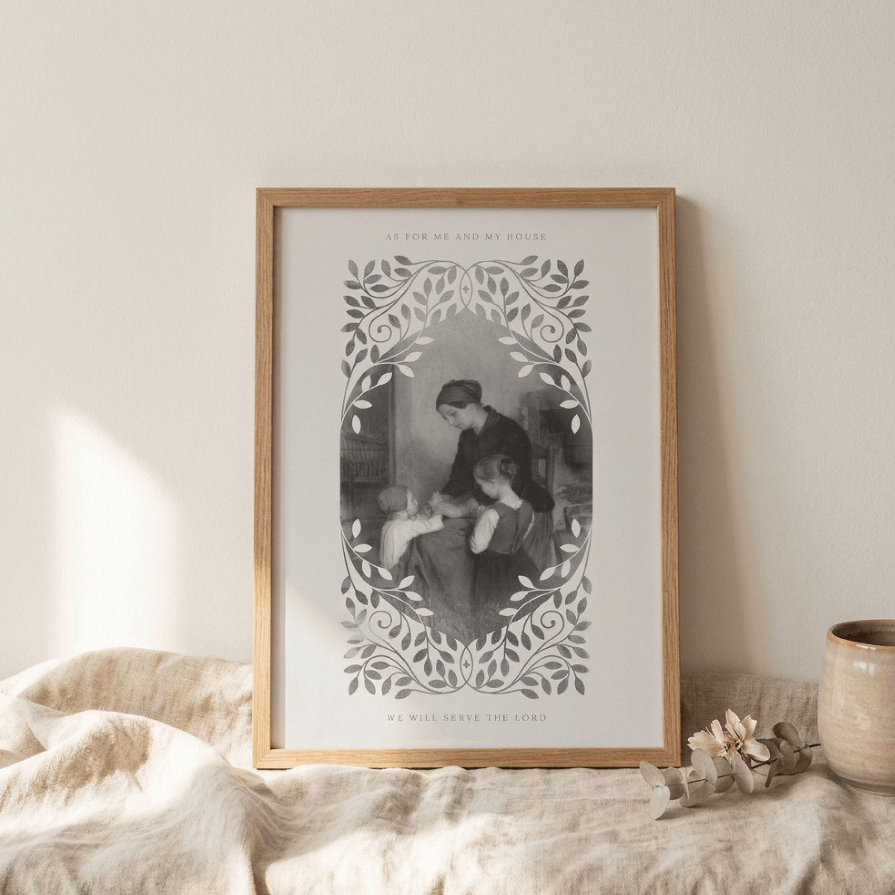 A framed black and white art print featuring a vintage illustration of a mother and child, surrounded by a decorative leafy border, displayed in a light wood frame on a draped beige fabric surface.
