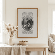 A framed black and white art print with a decorative leaf border hangs on a cream wall above a light wood console table with a vase of dried grasses and a bowl of nuts.