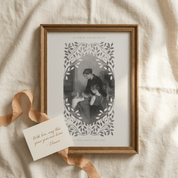A framed vintage-style art print featuring a monochrome illustration of a woman and child surrounded by ornate leafy scrollwork, styled on a cream linen fabric with a peach ribbon and a small card.