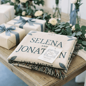 Gifts wrapped in paper with ribbons and a blanket with 'Selena and Jonathan' text on a wooden table.