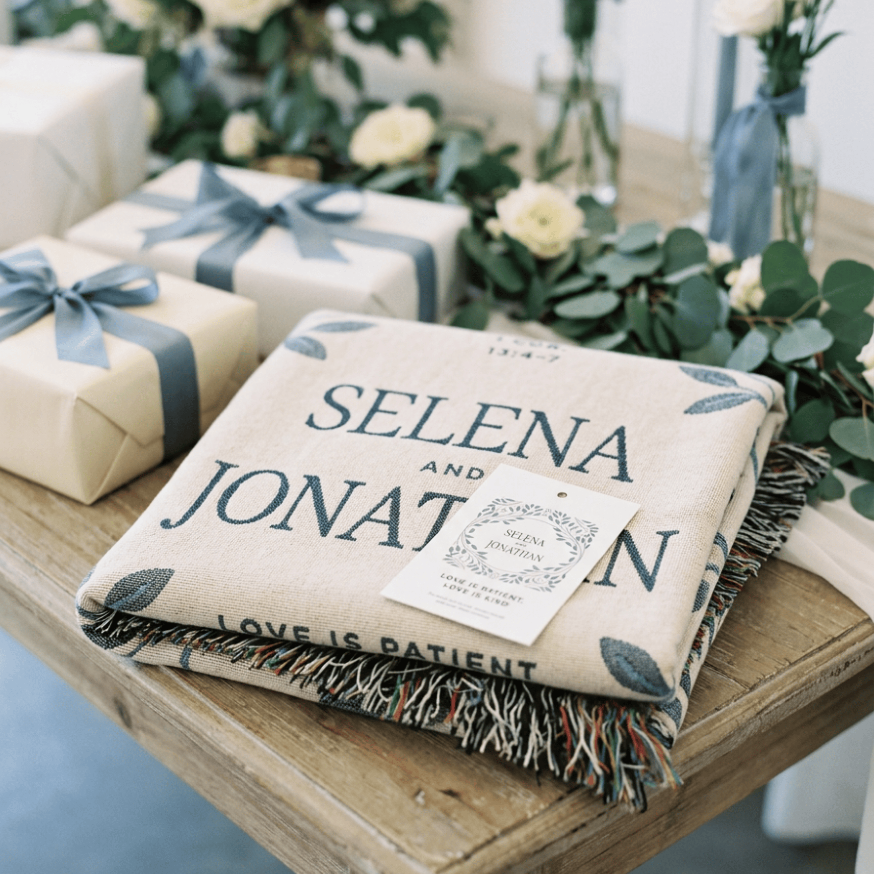 Gifts wrapped in paper with ribbons and a blanket with 'Selena and Jonathan' text on a wooden table.