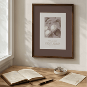 Framed artwork on a wall above a wooden desk with an open book and pen.