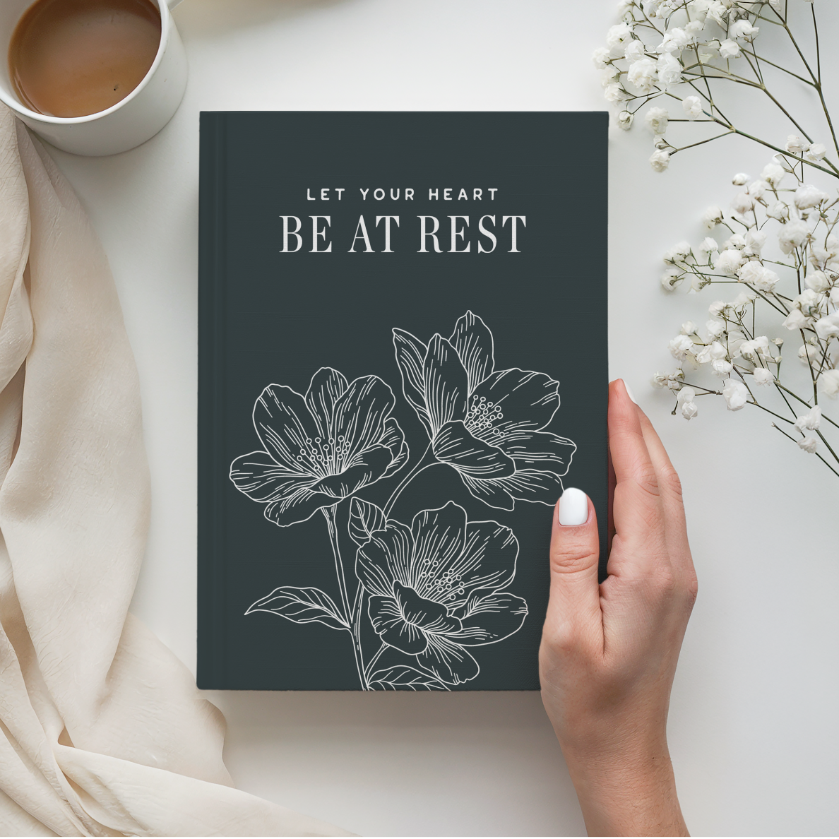Book titled 'Let Your Heart Be at Rest' with floral design held by a hand, next to a cup of coffee and white flowers.