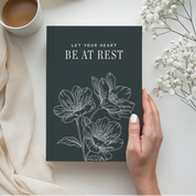 Book titled 'Let Your Heart Be at Rest' with floral design held by a hand, next to a cup of coffee and white flowers.