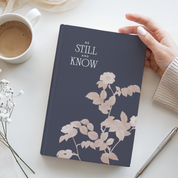 Notebook titled 'Be Still and Know' on a table with a cup of coffee and flowers.