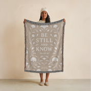 Cream-colored woven blanket with the phrase “Be still and know” from Psalm 46, made of 100% cotton with fringe; a peaceful Christian gift for nurseries or weddings.