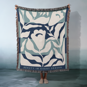 Floral patterned blanket with 'Beauty in Every Season' text held by a person against a gray background