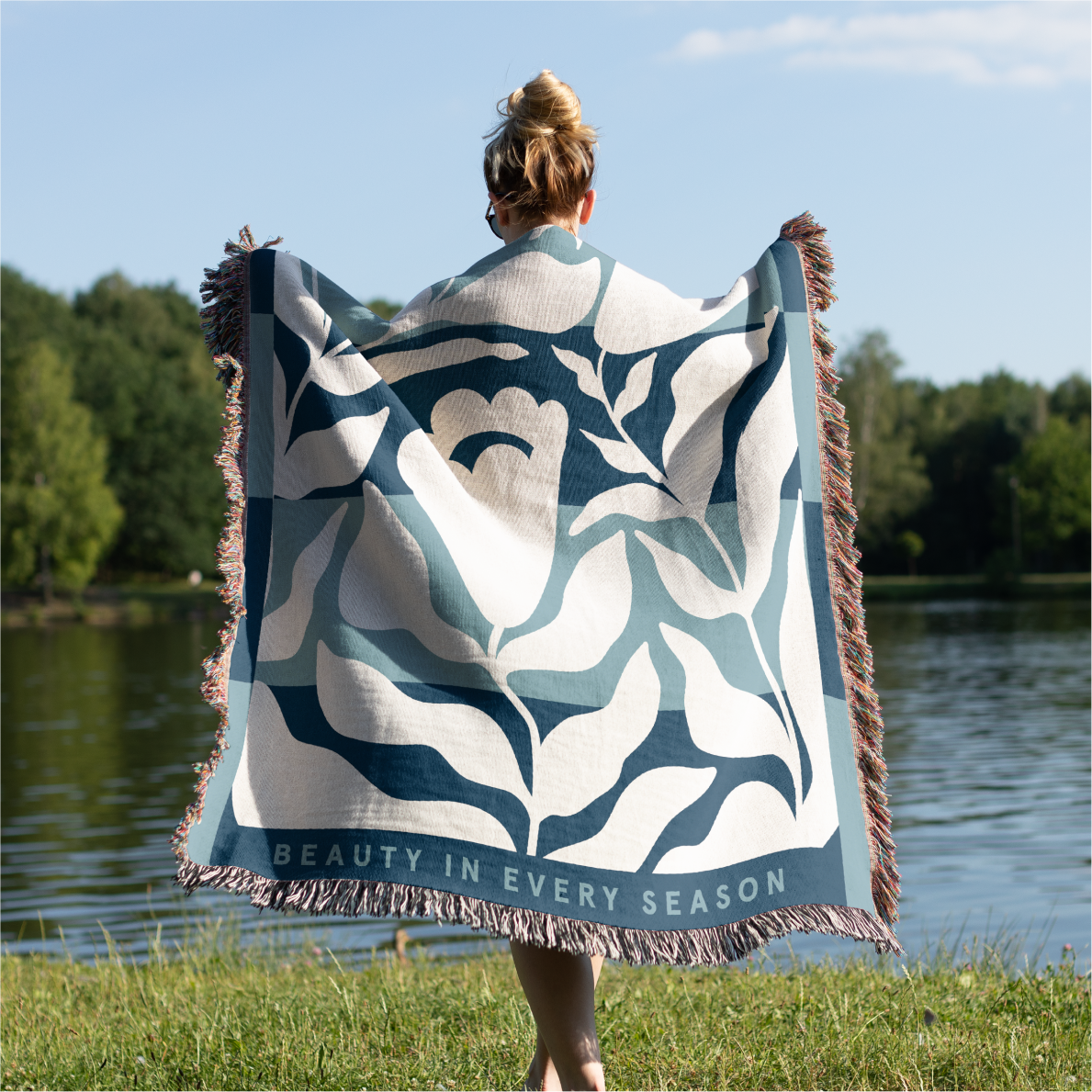 Person holding a decorative blanket with floral patterns and 'Beauty in Every Season' text by a body of water.