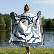 Person holding a decorative blanket with floral patterns and 'Beauty in Every Season' text by a body of water.