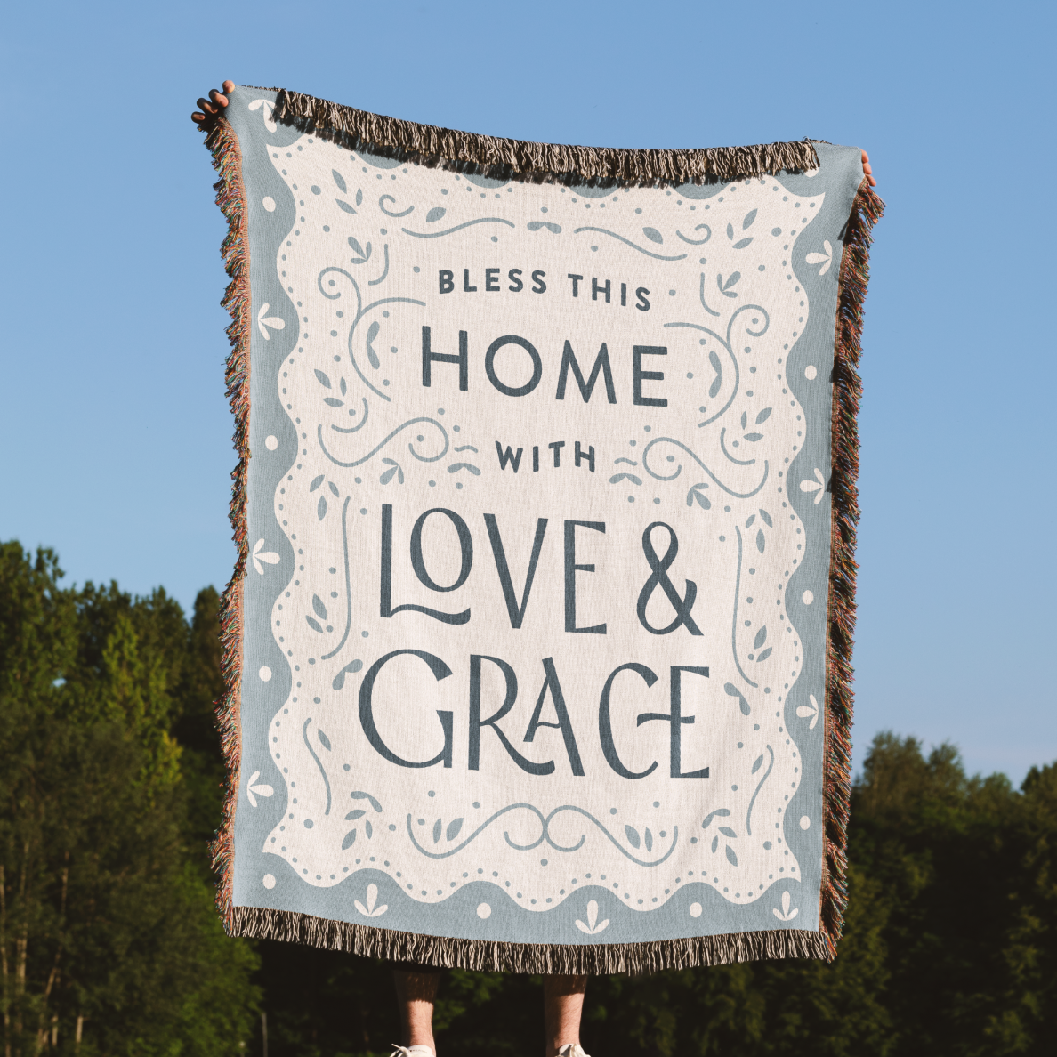Blanket with 'Bless this home with love & grace' text against a blue sky and trees background