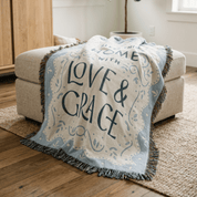 A cream and light blue throw blanket with fringe and the words “LOVE & GRACE” draped over a beige upholstered ottoman in a bright living room.