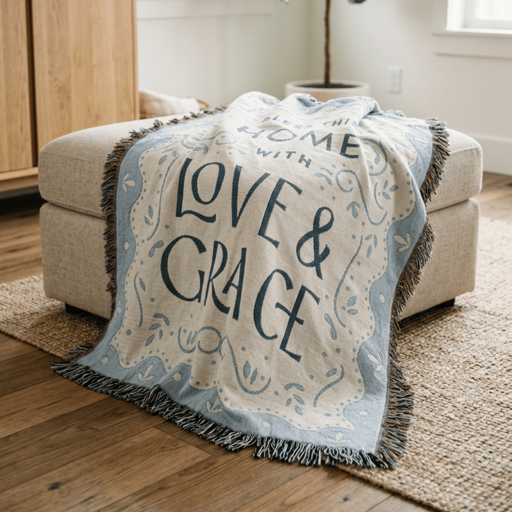A cream and light blue throw blanket with fringe and the words “LOVE & GRACE” draped over a beige upholstered ottoman in a bright living room.
