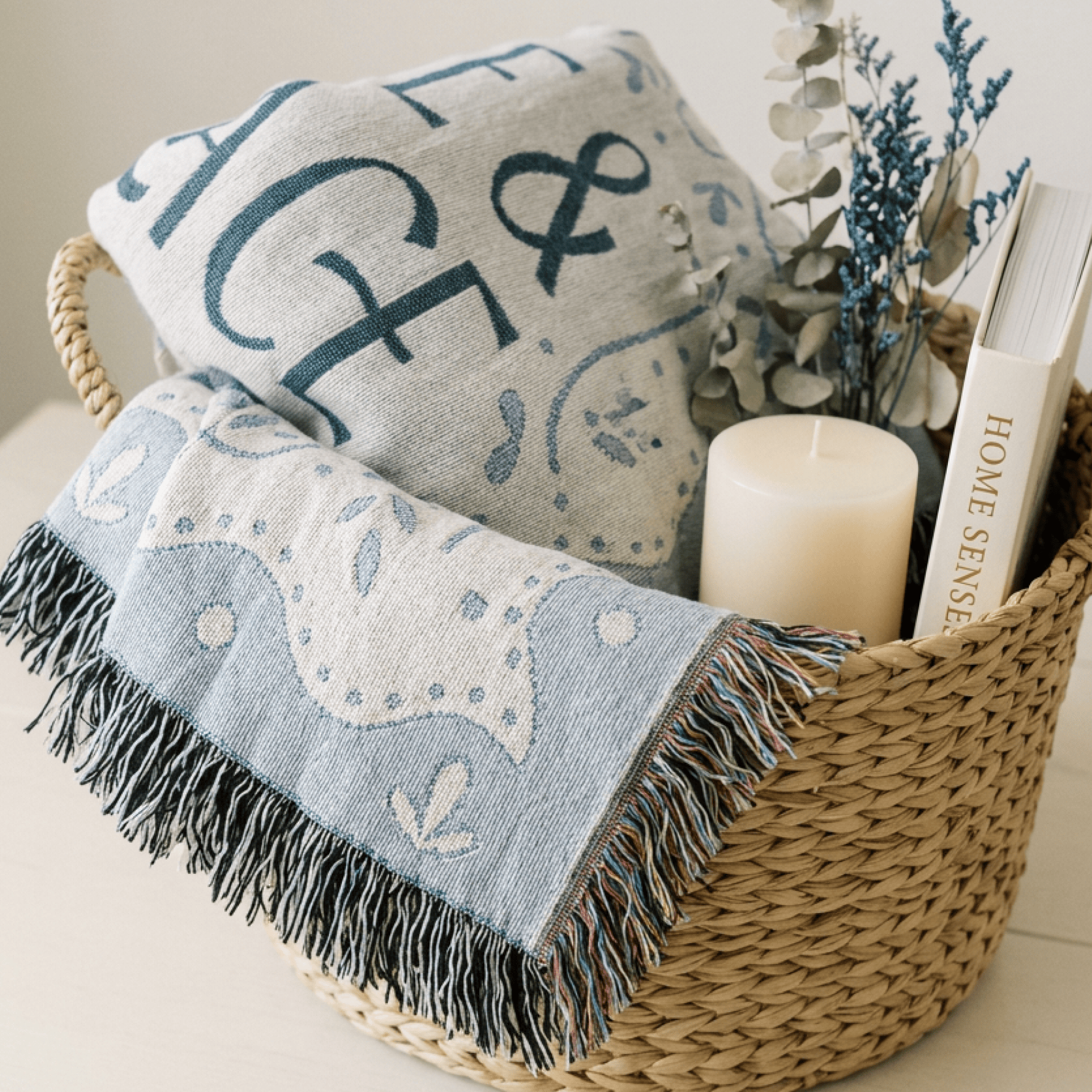 A woven basket holds two folded textile pieces with blue and white decorative patterns and fringe, styled with a candle, dried stems, and a book labeled HOME SENSE.