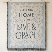 A square woven wall tapestry hanging from a wooden dowel against a textured off-white wall. The tapestry features the words 'BLESS THIS HOME WITH LOVE & GRACE' in dark blue-gray lettering, surrounded by a scalloped floral border.