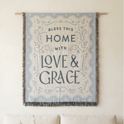 A decorative wall tapestry hanging from a wooden rod on a cream wall above a sofa. The tapestry has a light blue border with a cream center and the text 'Bless This Home With Love & Grace' in dark blue lettering.