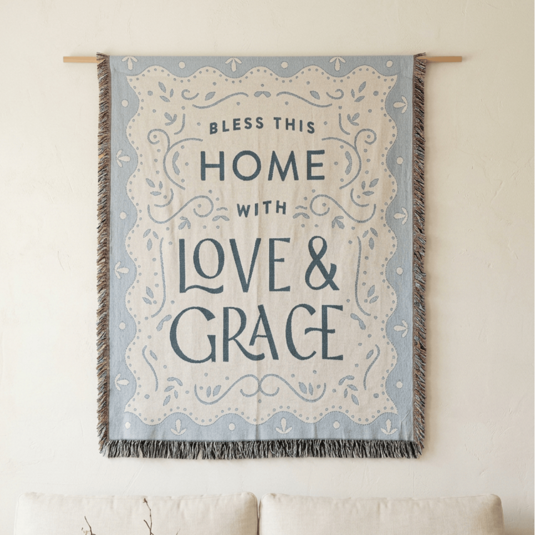A decorative wall tapestry hanging from a wooden rod on a cream wall above a sofa. The tapestry has a light blue border with a cream center and the text 'Bless This Home With Love & Grace' in dark blue lettering.