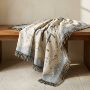 A woven throw blanket with fringe, draped over a wooden bench in a warm, sunlit room. The blanket has a cream base with a light blue border and abstract black and gray motifs.