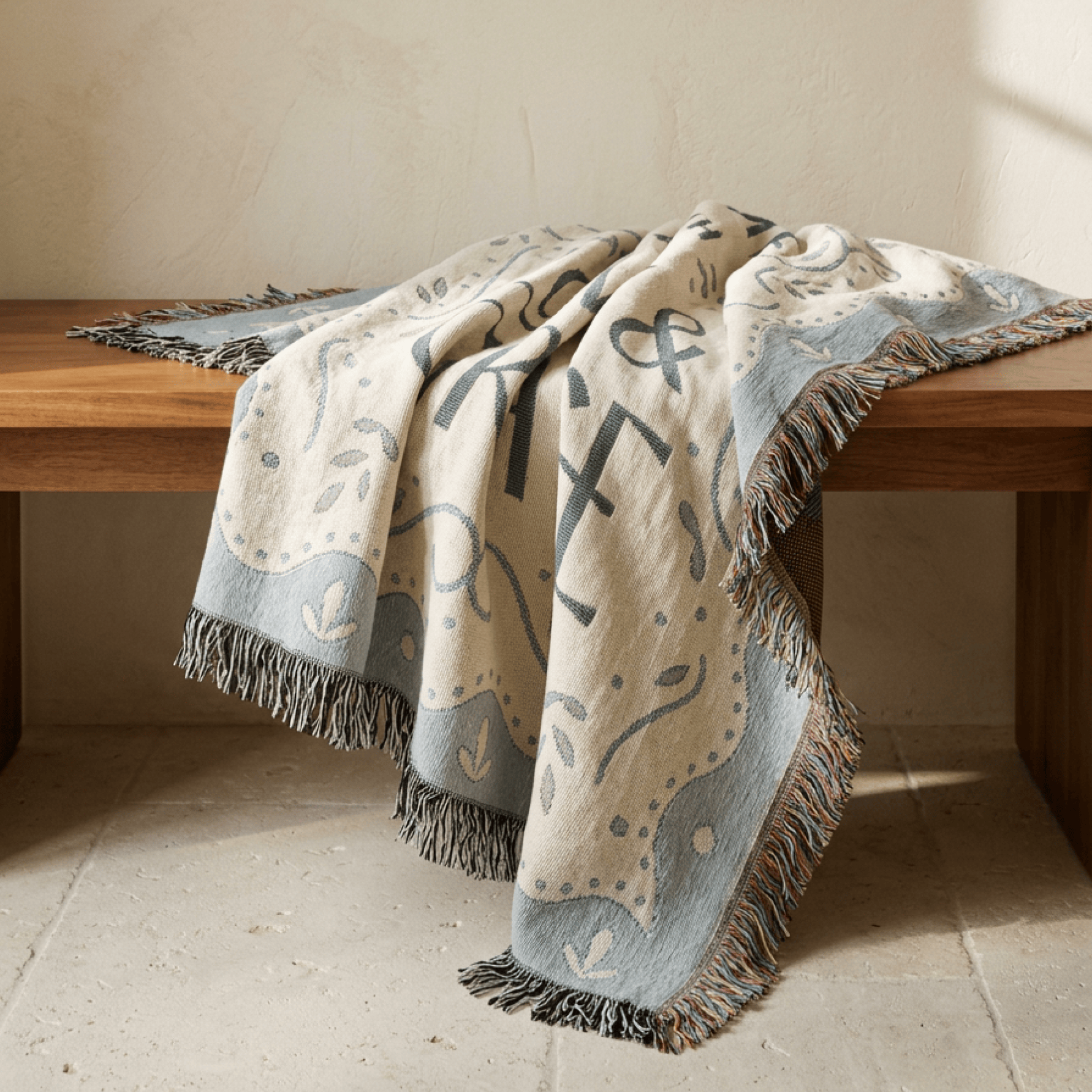 A woven throw blanket with fringe, draped over a wooden bench in a warm, sunlit room. The blanket has a cream base with a light blue border and abstract black and gray motifs.