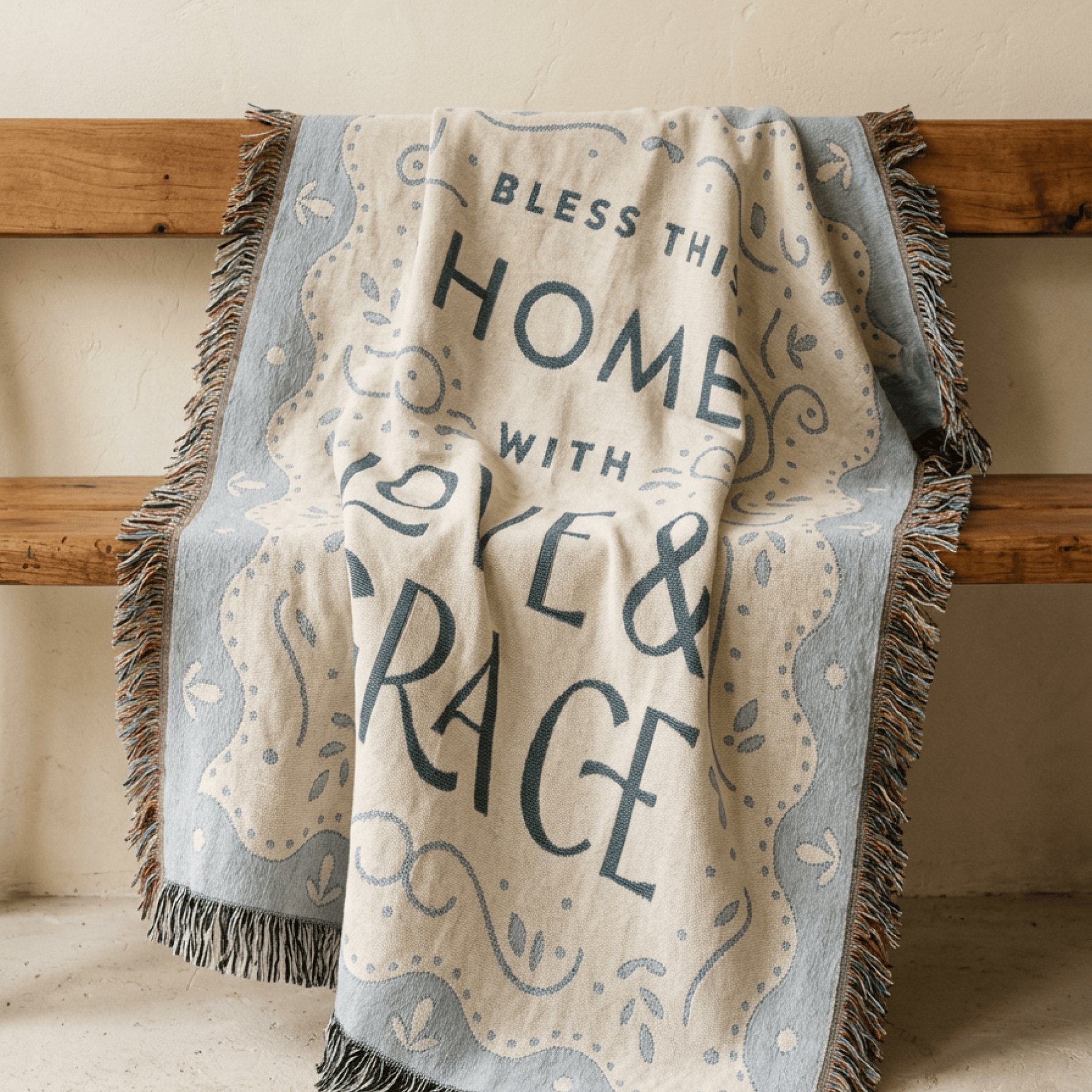 A fringed throw blanket draped over a wooden bench, featuring the printed text 'Bless This Home With Love & Grace' in dark blue lettering on a cream background with a light blue decorative border.