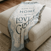 A beige woven throw blanket draped over a matching beige upholstered ottoman, featuring the phrase “Bless This Home With Love & Good” in dark lettering with decorative scrollwork and fringe edges.