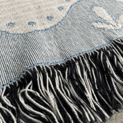 Close-up of a woven textile with a light blue and off-white patterned surface and a black-and-white fringe along the bottom edge.