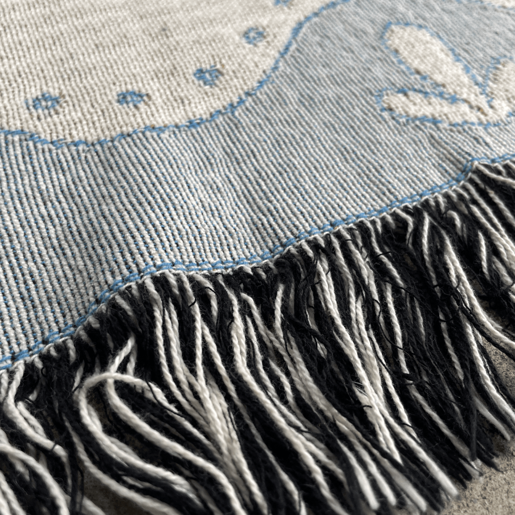 Close-up of a woven textile with a light blue and off-white patterned surface and a black-and-white fringe along the bottom edge.