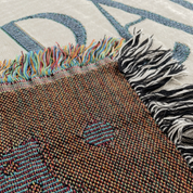 Close-up of a woven textile with multicolored fringe, showing a brown textured section with small blue and red woven details, laid over a light cream background with large blue lettering.