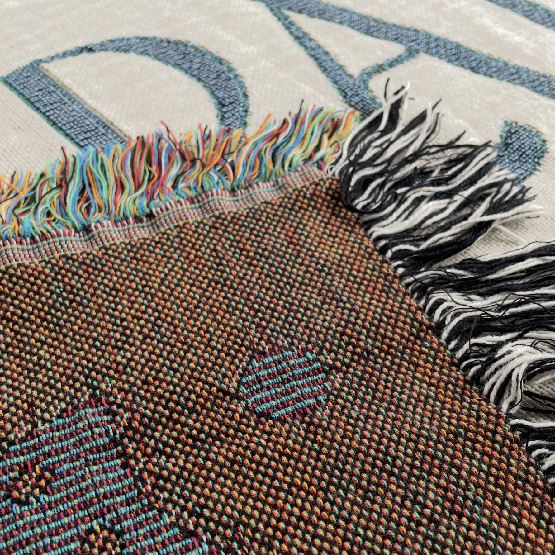 Close-up of a woven textile with multicolored fringe, showing a brown textured section with small blue and red woven details, laid over a light cream background with large blue lettering.