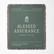 A square woven throw blanket with fringed edges, featuring a sage green background and white text that reads 'BLESSED ASSURANCE' with smaller scripture text around it. The design is centered on a light gray background.