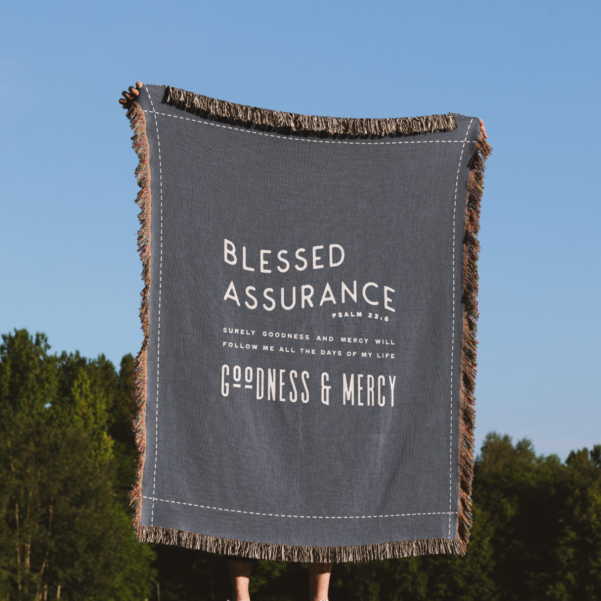 Gray blanket with 'Blessed Assurance' text against a blue sky and green trees background