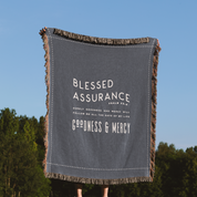 Gray blanket with 'Blessed Assurance' text against a blue sky and green trees background