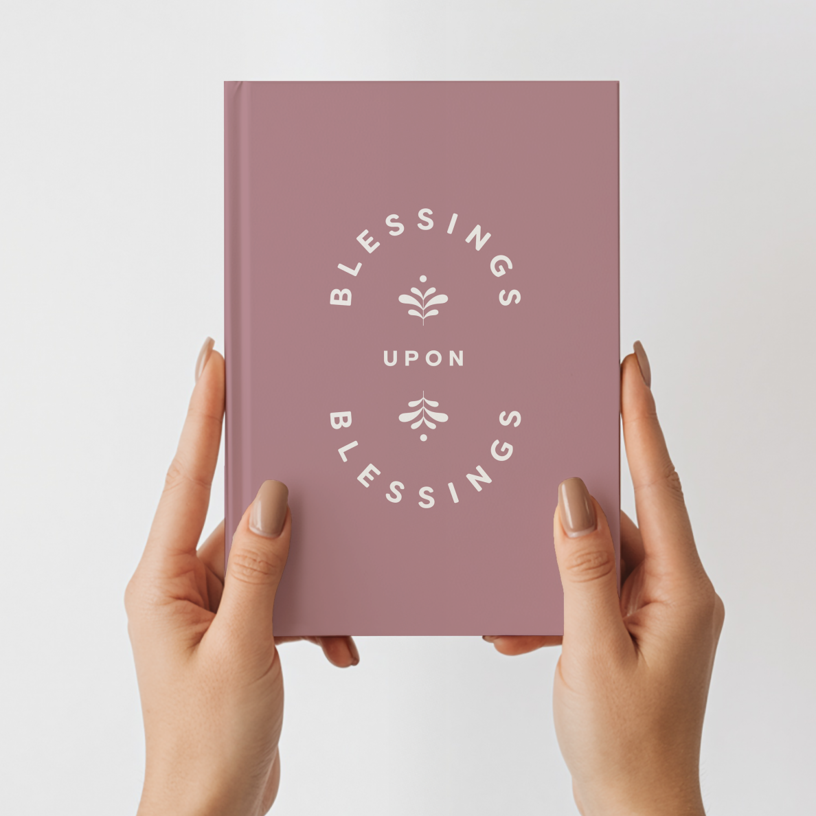 Pink notebook titled 'Blessings Upon Blessings' held by hands against a light gray background