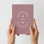 Pink notebook titled 'Blessings Upon Blessings' held by hands against a light gray background