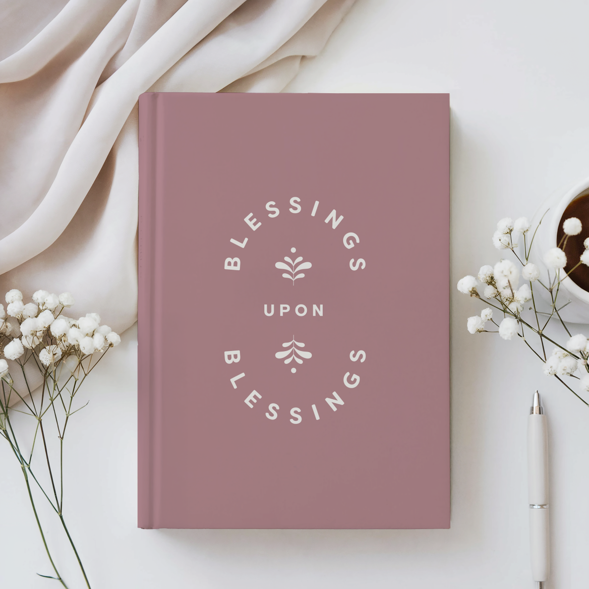 Pink book titled 'Blessings Upon Blessings' on a white surface with flowers and a pen.
