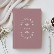 Pink book titled 'Blessings Upon Blessings' on a white surface with flowers and a pen.