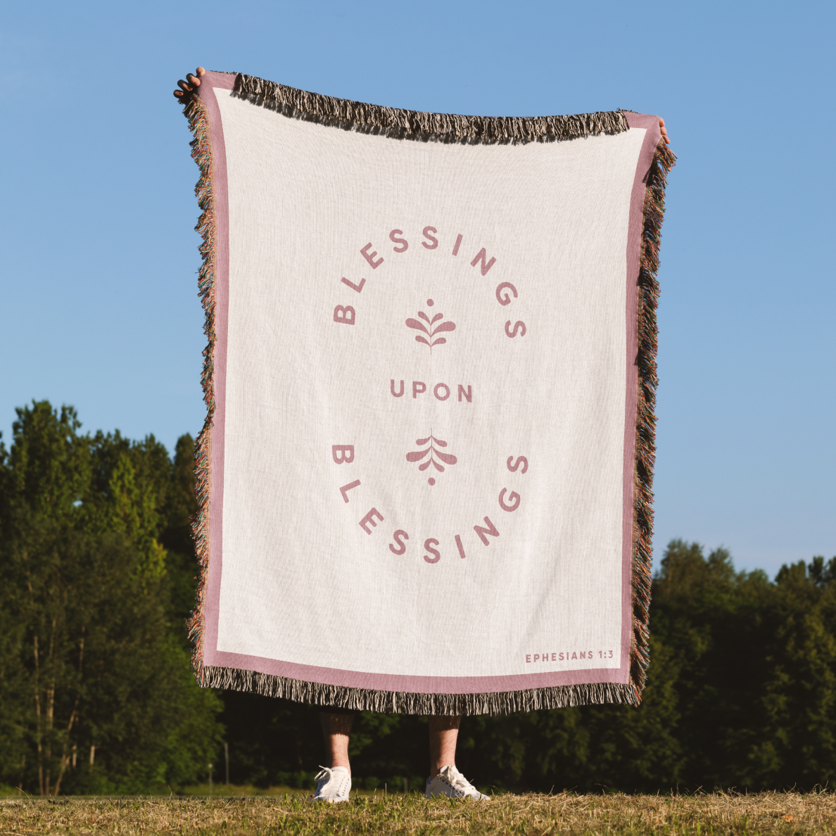 Blanket with 'Blessings Upon Blessings' text in a circular design on a grassy field with trees and blue sky.
