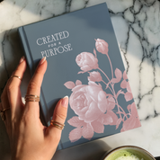 Book titled 'Created for a Purpose' with floral design held by a hand on a marble surface.
