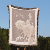 Blanket with floral design and 'Do Everything in Love' text against a blue sky and green trees background