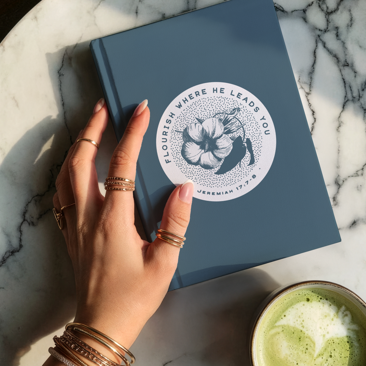 Hand holding a blue book with a decorative cover on a marble surface, next to a cup of green tea.