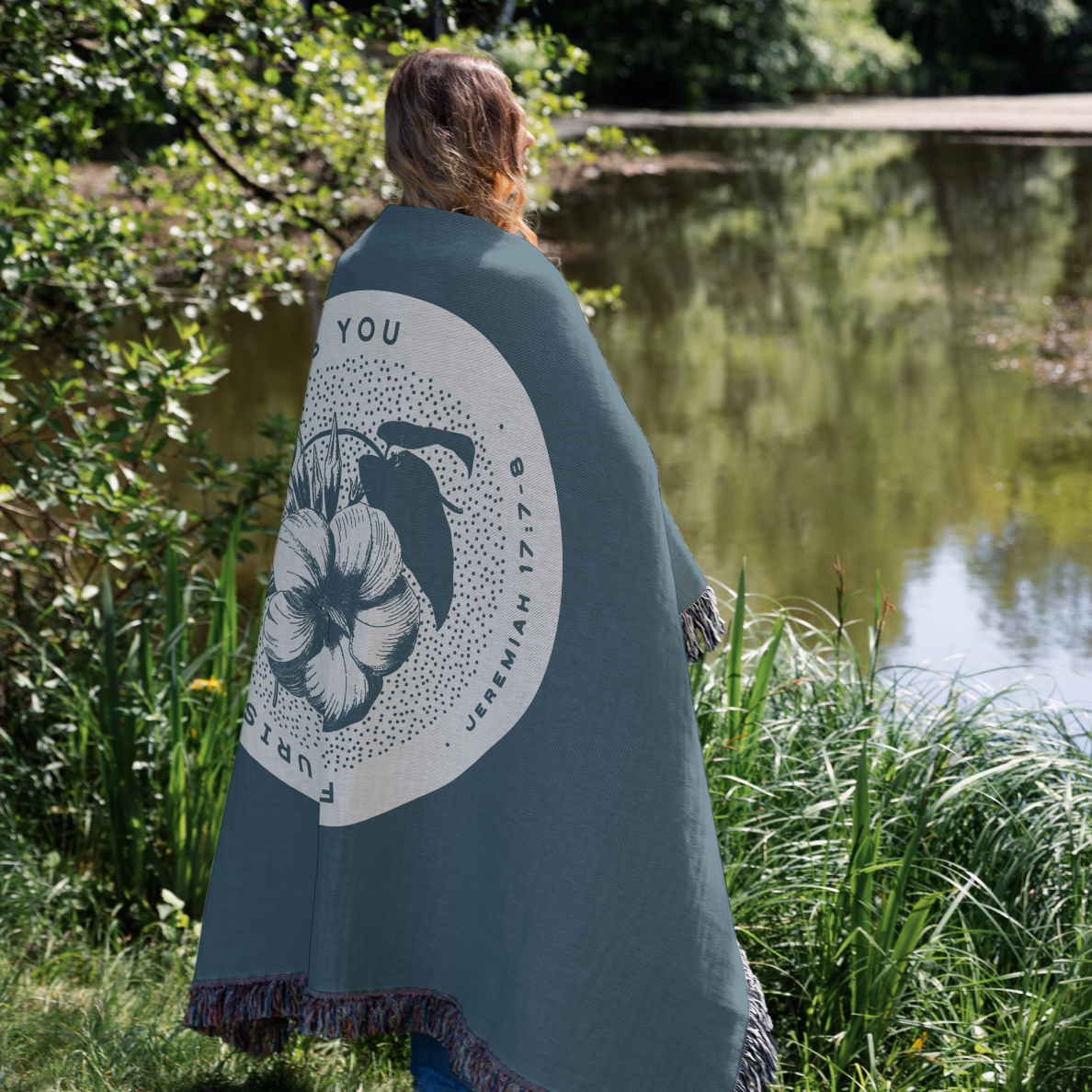 Person wrapped in a teal blanket with a floral design and text, standing by a pond.