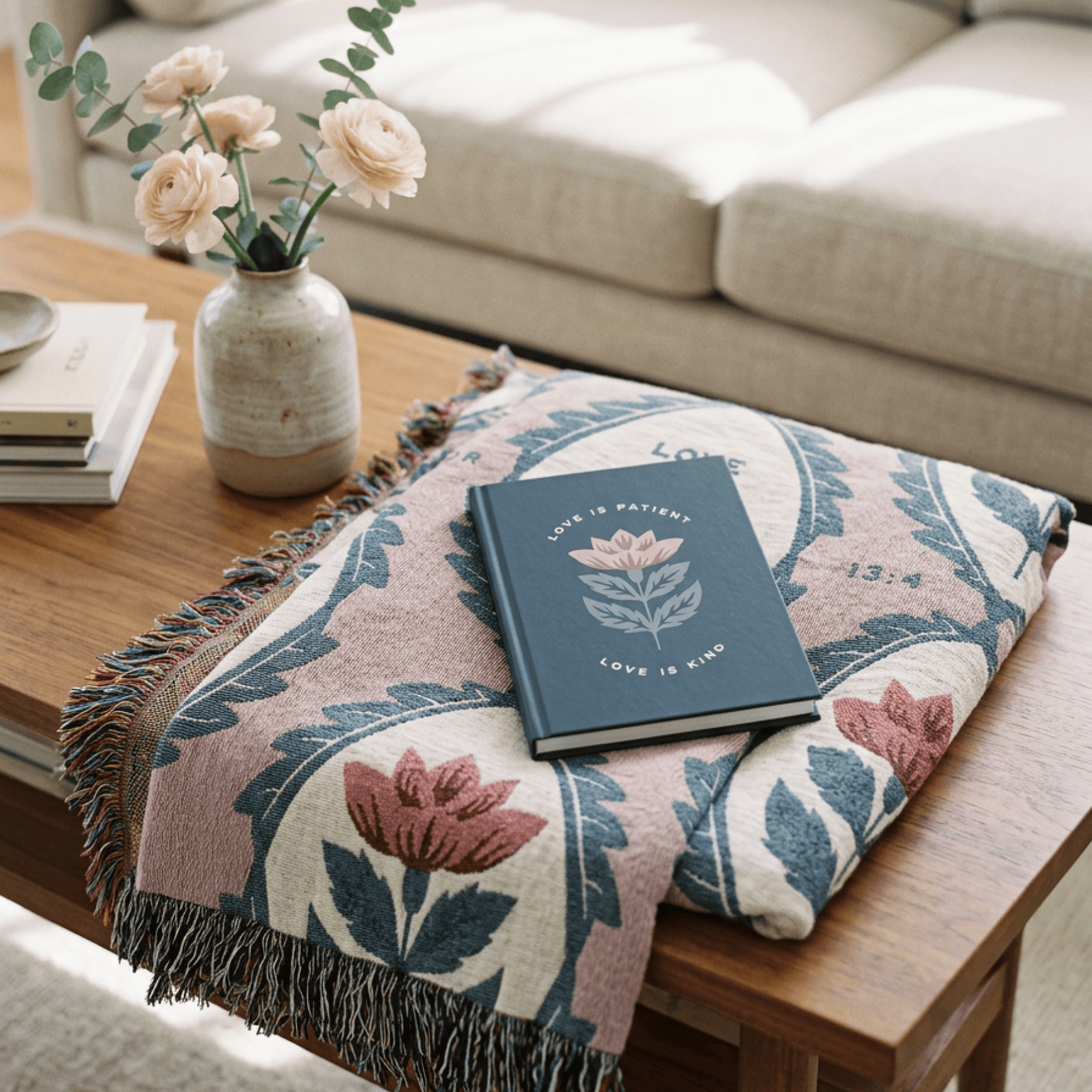 A folded throw blanket with a floral pattern in dusty pink, slate blue, and cream sits on a wooden table in a living room setting, styled with a navy blue book and a small vase of flowers.