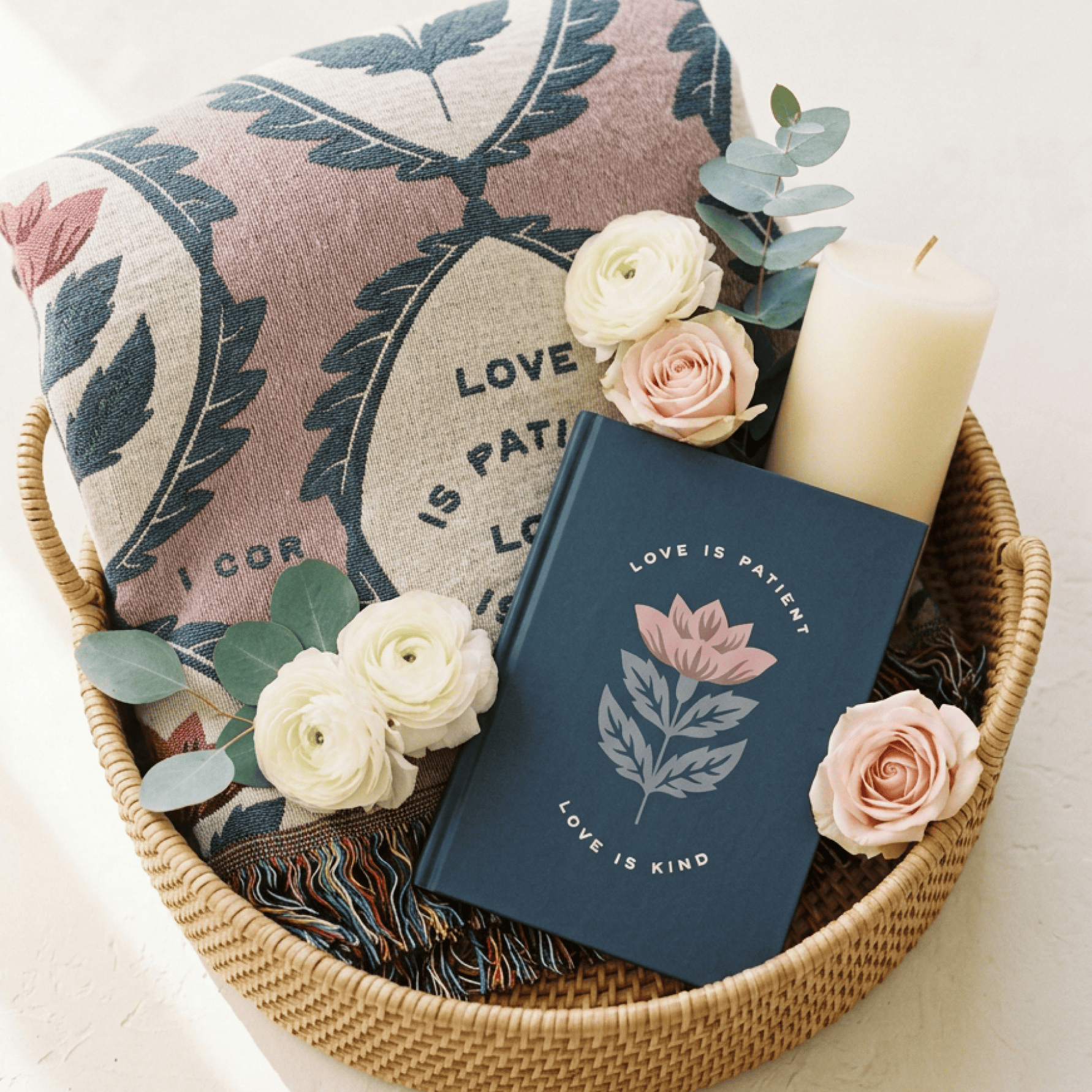 A styled gift basket arrangement featuring a navy blue journal with a pink lotus flower and the words 'LOVE IS PATIENCE LOVE IS KIND', a patterned throw blanket, cream and blush roses, green eucalyptus leaves, and a white pillar candle in a woven wicker basket.